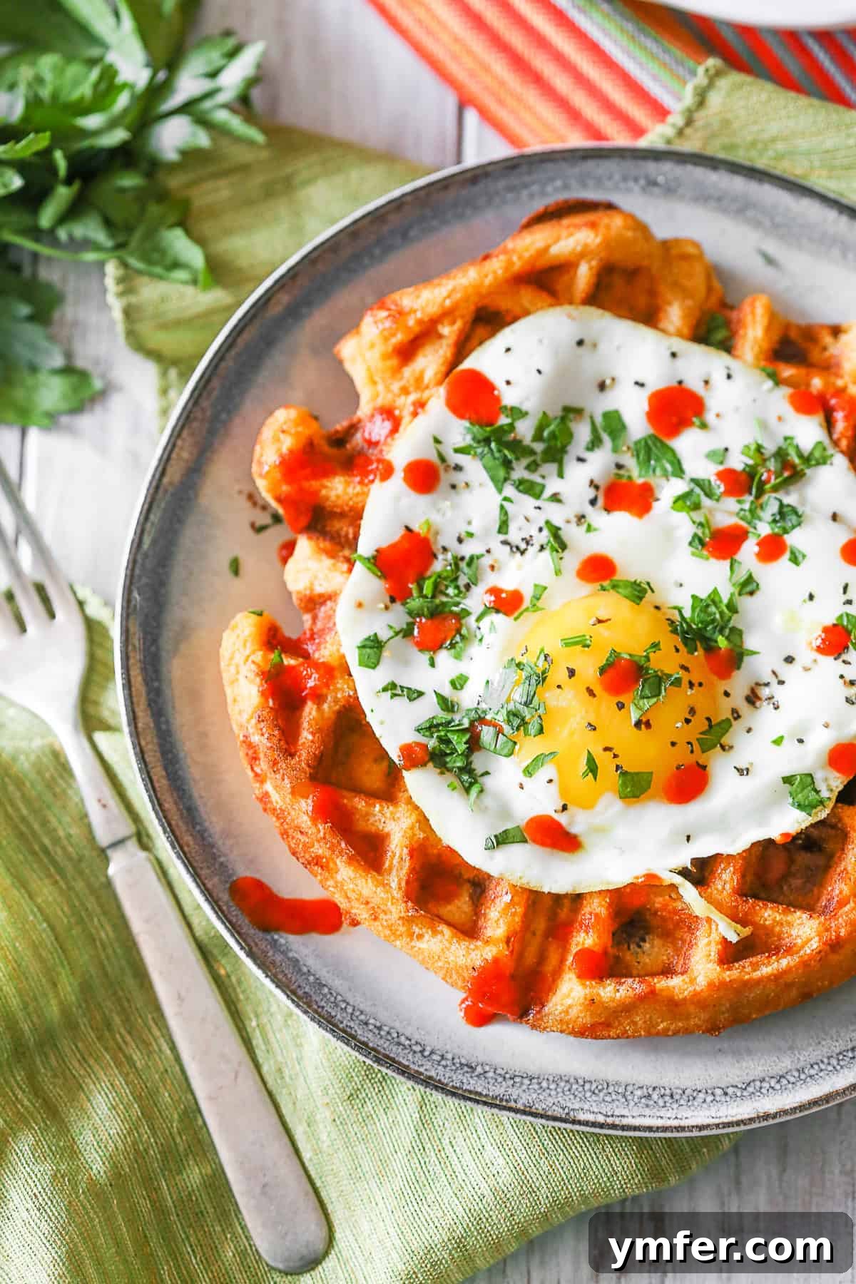 Savor the unique taste of Cheddar Sausage Cornmeal Waffles, infused with sriracha, cheddar, and breakfast sausage, perfect with a fried egg. Cheddar Sausage Cornmeal Waffles topped with cilantro and sriracha, served with a fried egg.