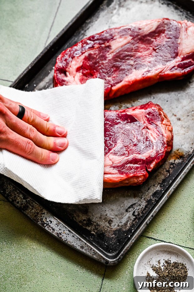 Perfect Pan-Seared Ribeye 4 Hands blotting excess moisture from a raw ribeye steak with a paper towel, a crucial step for achieving a perfect sear.