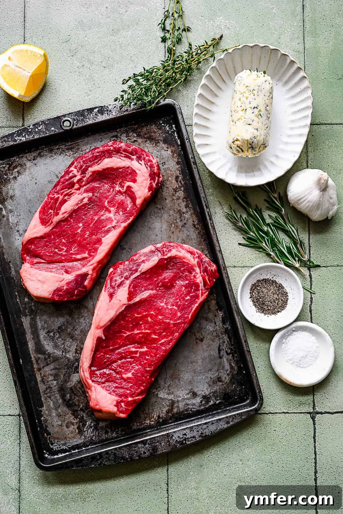Perfect Pan-Seared Ribeye 3 Raw ribeye steak, fresh seasonings, herbs, and compound butter arranged on a rustic wooden table, ready for cooking.