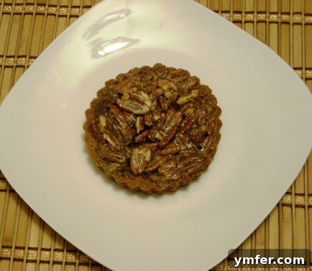 Espresso-Kissed Chocolate Pecan Bites 3 A single Mini Chocolate Espresso Pecan Tart, showcasing its rich brown filling and delicate pecan topping, artfully placed on a small white plate.