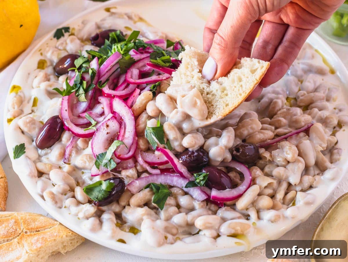 Piyaz Zesty Turkish White Bean Salad 6 A serving of Piyaz on a white plate, with a hand holding a piece of pita bread ready to scoop